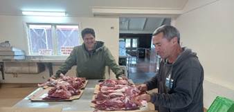 A couple of men standing around a table with meat

Description automatically generated